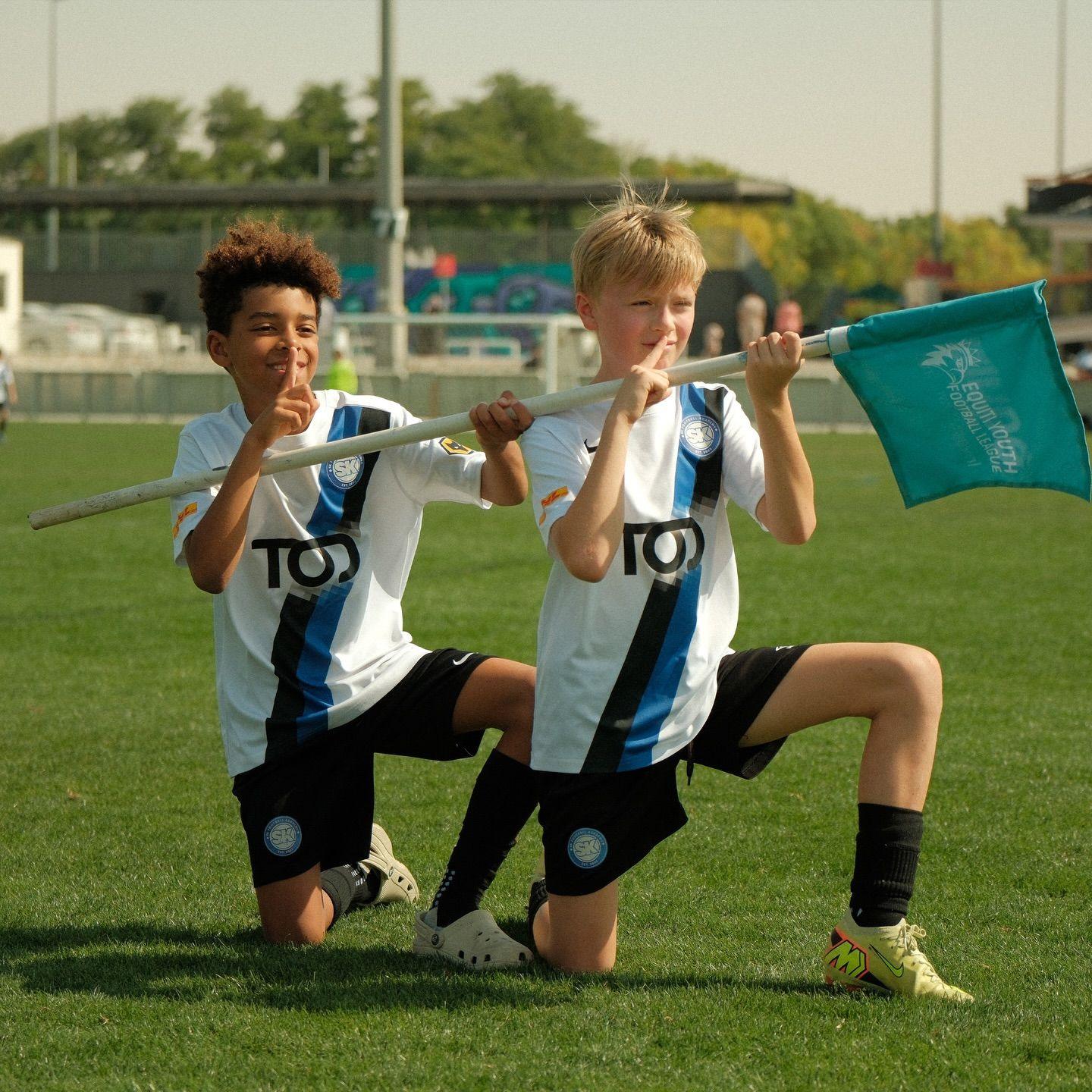 Two EYFL teammates kneel on the pitch holding a corner flag